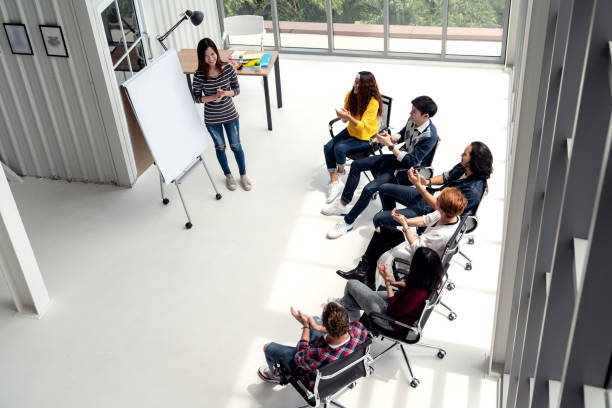 Young asian businesswoman explain idea to group of creative diverse team at modern office. Wide top view of manager standing against multiethnic people. Audience applauding speaker after presentation.